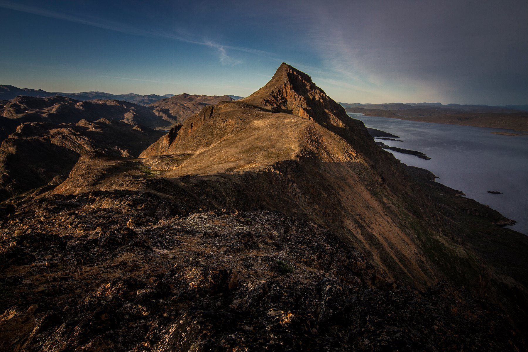 Rocky mountain ridge and peak overlooking a wide body of water under a clear sky at dusk; brown, gold and blue tones, dramatic and calm